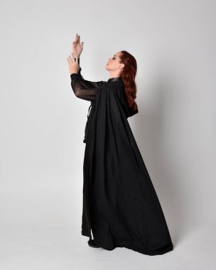 Kızıl saçlı bir kadının fantezi portresi. Uzun siyah pelerinli koyu deri bir suikastçı kostümü giyiyor. Stüdyo arka planında tam boy ayakta poz.