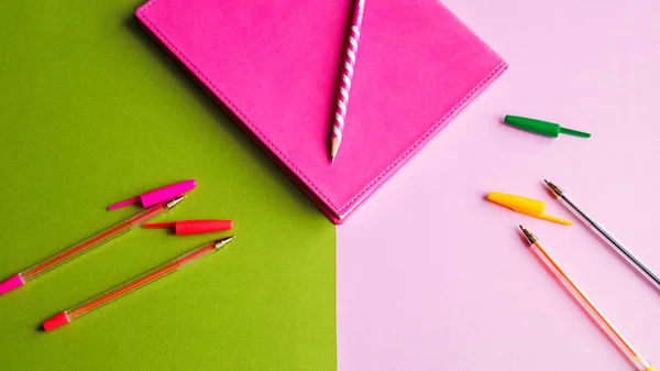 colorful pens and pencil on pink notebook on green and pink background, education concept