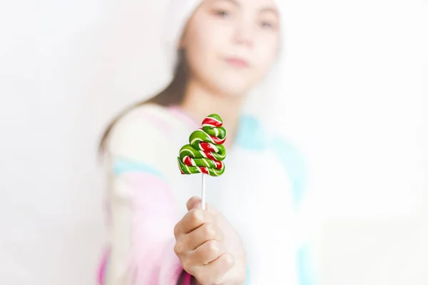 Joven linda chica en sombrero de Santa sobre fondo blanco sostiene una piruleta en la forma de un árbol de Navidad en su mano. Fondo navideño festivo — Foto de Stock