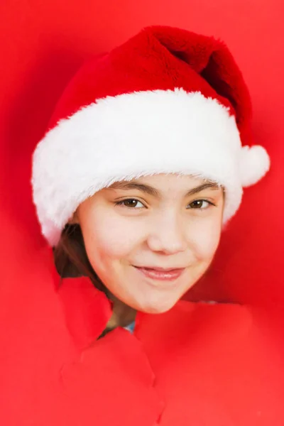 Pohled na dívku v Santově klobouku skrz díru v červeném papíru. Usmívající se dívka udělá díru v novinách. Slavnostní vánoční pozadí — Stock fotografie