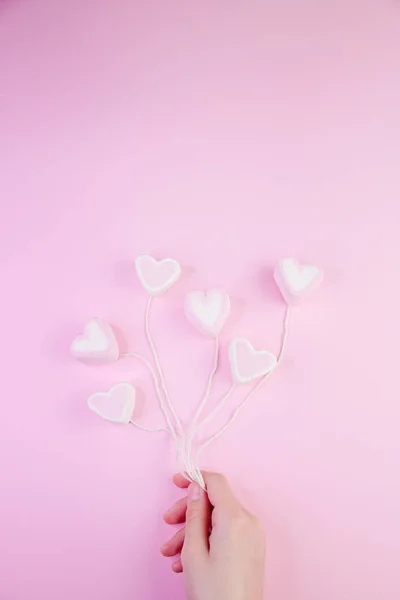 Lo sfondo di San Valentino pastello è fatto di soufflé di marshmallow nella forma di cuori su una corda che tiene una mano su uno sfondo rosa. Concetto di San Valentino con spazio di copia — Foto Stock