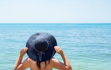 Islak saçlı ve şapkalı bir kadın sırtı kameraya dönük duruyor, şapkasını tutuyor ve denize bakıyor..