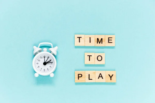 Flatlay concept time to play, white alarm and letters wooden cube on blue background. Selective focus.