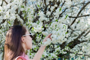 Okul çağındaki genç bir kızın saçları çiçek açan bir elma ağacının arka planında poz veriyor ve sıcak havada dalı tutuyor. Seçici odak.