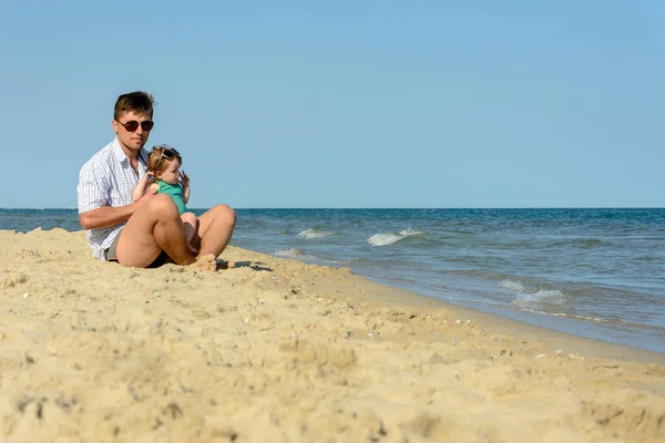 Un père avec une petite fille est assis sur la plage à l'arrière-plan de la mer — Photo