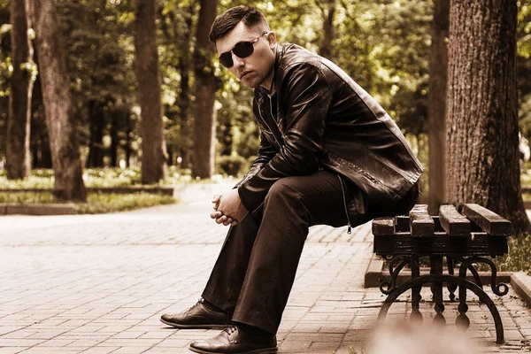 Güneş gözlüğü ve bir deri ceket bir bankta bir parkta oturan genç adam — Stok fotoğraf