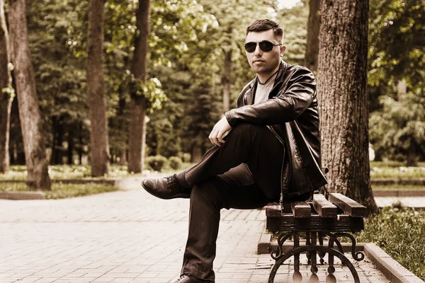 Молодий чоловік сидить у парку на лавці в сонцезахисних окулярах і шкіряній куртці — стокове фото