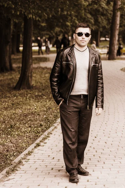 Young handsome man standing in a park in sunglasses and a leather jacket — Stock Photo, Image