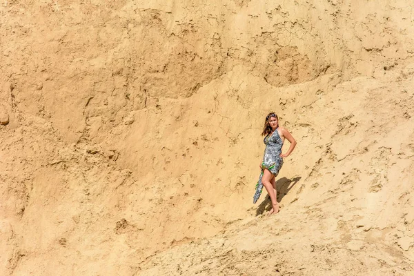 Une belle femme se tient dans une robe et des lunettes de soleil contre une montagne de sable — Photo