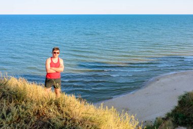 Kırmızı gömlekli genç bir adam denizin arka planında bir tepede oturuyor.