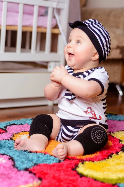 Um bebê bonito em uma camisa listrada e chapéus sentados no tapete na sala — Fotografia de Stock