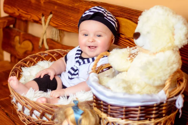Um lindo bebê em um terno de marinheiros senta-se em uma cesta ao lado de um urso de pelúcia — Fotografia de Stock