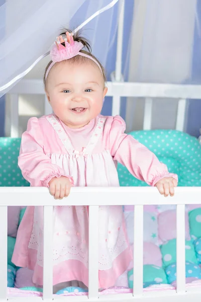 Pequena menina bonita em um vestido rosa ri em uma cama de bebê — Fotografia de Stock