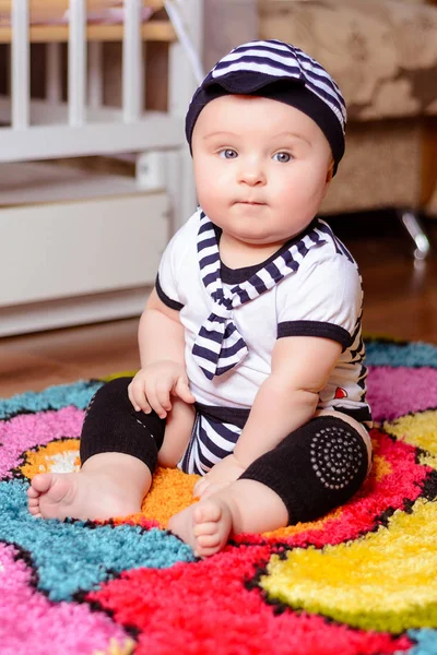 Un bonito bebé con una camisa a rayas y sombreros sentados en la alfombra de la habitación — Foto de Stock