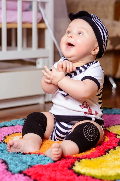 Um bebê bonito em uma camisa listrada e chapéus sentados no tapete na sala — Fotografia de Stock