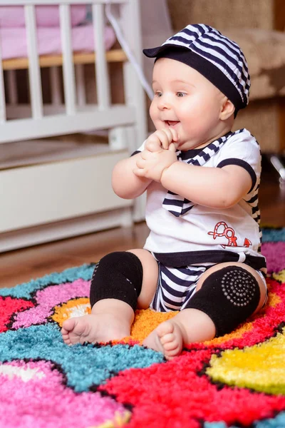 Um bebê bonito em uma camisa listrada e chapéus sentados no tapete na sala — Fotografia de Stock