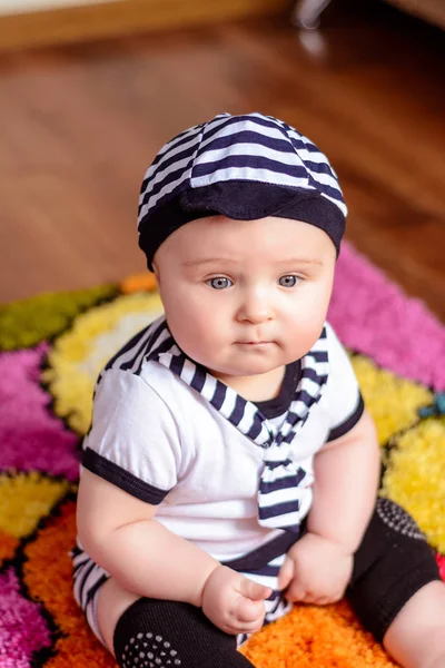 Um bebê bonito em uma camisa listrada e chapéus sentados no tapete na sala — Fotografia de Stock