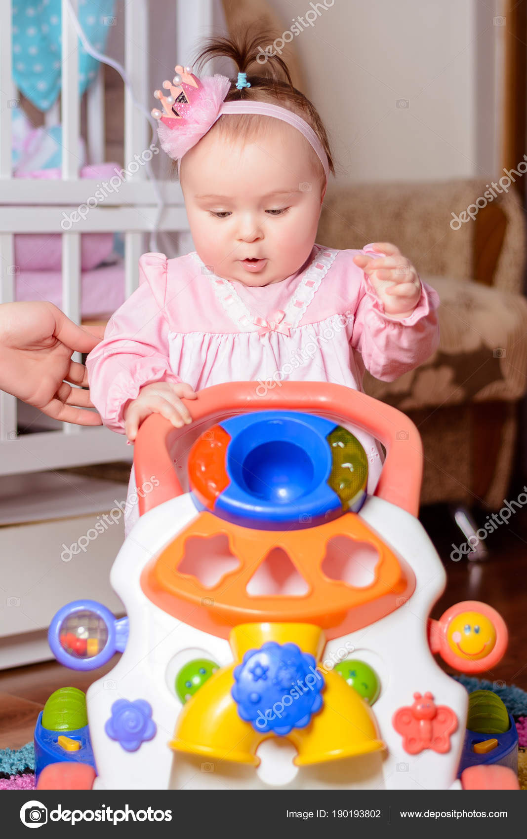 A Little Girl In A Pink Dress Is Playing A Toy Car Near The Crib