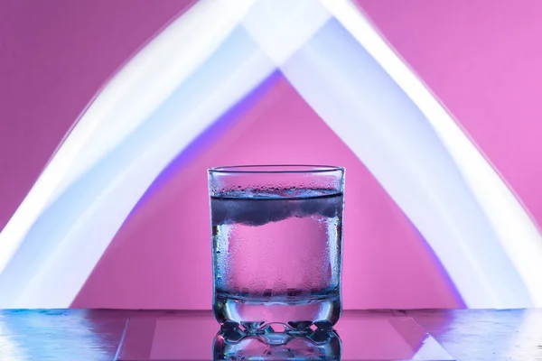 Líquido con hielo en un vaso sobre una superficie reflectante sobre un fondo rojizo —  Fotos de Stock
