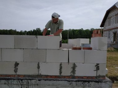 Usta inşaat işi yapıyor, yani, gaz bloklarından oluşan bir duvar inşa ediyor. Aletler ve yapıştırıcı harç ile, arka planda gökyüzü ve orman..