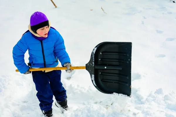 Lächelnder Junge trägt Schnee auf einer Schaufel, Kind säubert nach Schneefall den Hof. — Stockfoto