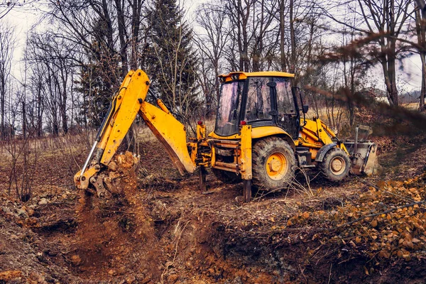 Uma escavadora industrial enferrujada amarela com um balde no chão, cavando o chão em um canteiro de obras em uma floresta de lagoas . — Fotografia de Stock