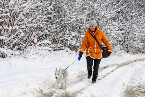 Piękna kobieta w pomarańczowej kurtce z husky szczeniaczkiem biegnącym przez leśną drogę. — Zdjęcie stockowe