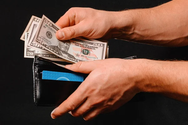 Carteira preta com dólares em mãos de homem sobre fundo preto . — Fotografia de Stock