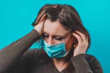 Uzun saçlı, koruyucu tıbbi maskeli, baş ağrısı ve ağlayan gözleri olan genç bir kadın. 2020