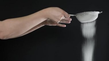 Mutfakta, un ya da şekere benzer beyaz tozu siyah arka planda metal elek yardımıyla elemekte olan kadın 2020.