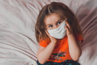 Karantina sırasında Coronavirus 2021 ile yatakta oturan maskeli küçük kız mutlu bir şekilde görünüyor.