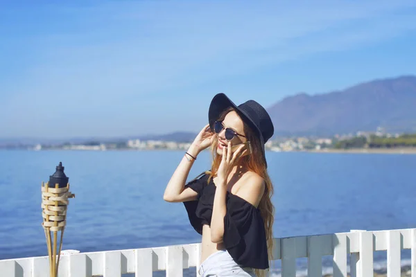 女人在电话里聊天 可爱的女孩微笑 海的背景 连接和沟通的概念在旅途中 穿帽子的女孩 — 图库照片