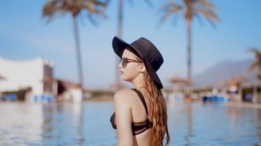 Sarışın kız sigara elektronik sigara yakınındaki bir şapka ve güneş gözlüğü havuzunda sweaming. Beackgroung palm, güneş, Havuzu. Genç güzel kadın (vaping) e-sigara bir plajda. Tatil. Turizm.