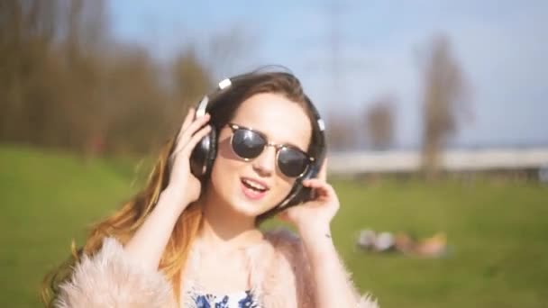 女孩在蓝牙耳机 唱歌和舞蹈外 一个甜美的女孩 长头发 太阳镜在公园里跳舞和汗水 对蓝天 — 图库视频影像