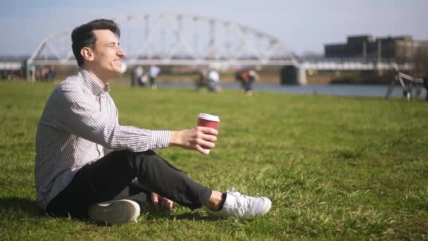 坐在公园里的人在草地上 享受着阳光 喝着咖啡 一个男人坐在河边 人们围着背景走 幸运日 桥的背景 — 图库视频影像