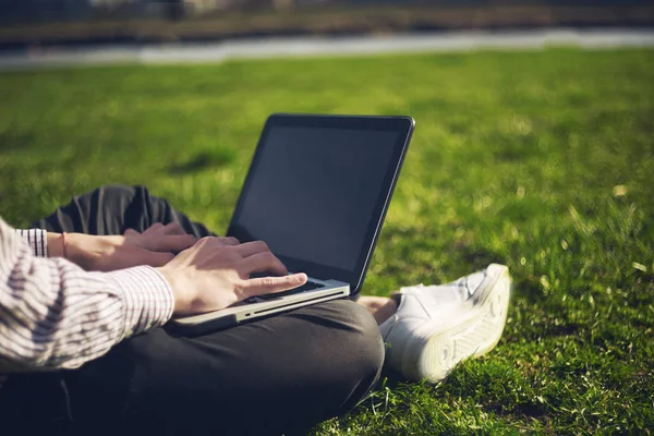 Young man in the park sitting on the grass with a laptop. Man using and typing laptop computer in summer grass. Man with laptop outdoor. Cropped shot of man using laptop with blank screen while sitting on green grass. Hipster filter.