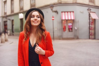 Eski şehir sokakta önünde poz şık hippi kız. Trendy genç kadın modeli. Sokak moda kavramı, şapka ve kırmızı ceket giyiyor. Turist gülümseme, güneşli bir gün. Uzun kıvırcık saçları zarif kız