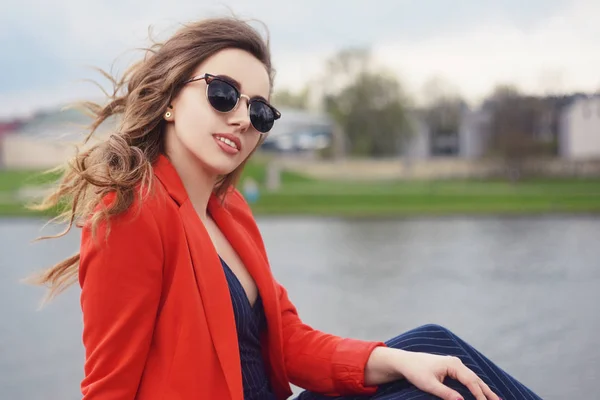 美しい女の子川沿いでサングラスをかけている 女の子の桟橋に座って 川を見て 晴れた日には屋外で美しい若い女の子 ファッションモデル旅行 女の子とともに長い髪で赤いジャケット — ストック写真