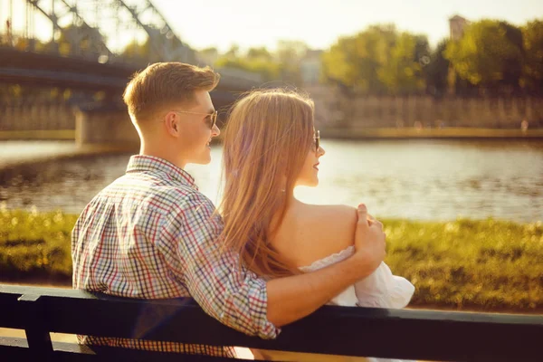 川の近くの公園のベンチに座っている美しい若いカップル 川沿いのベンチにロマンチックなカップルはデートがあります 笑顔観光 サングラス笑顔 夏のカップル — ストック写真