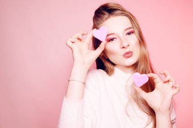 The blonde holds a sweet pink heart near her face, on a pink bac