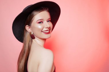 Büyük şapkalı, bordo makyajlı, kırmızı dudaklı ve bordo elbiseli güzel bir sarışın. Burgundy küpeli bir kız, bir mankenin portresi. Parlak kırmızı makyajlı moda fotoğrafı, kırmızı arka planda model. Gülümse..