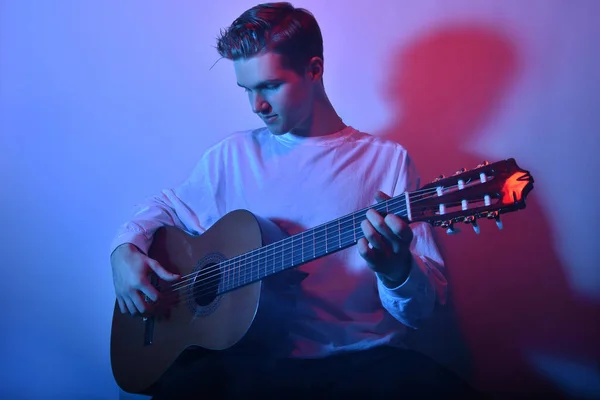 A man plays an acoustic guitar in neon red-blue light. A man learns to play the guitar, music, hobbies.