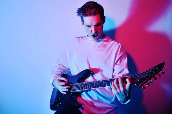 A man plays the electric guitar in neon light, red blue light. Hobbies, music, club. A man enjoys playing the guitar, screaming, vivid emotions.