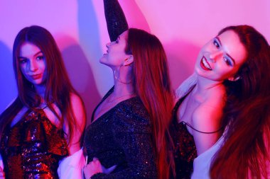Üç güzel kız neon ışıkta dans ediyor. Parlak elbiseli kızlarla inanılmaz eğlenceli bir parti, bayramı kutluyorlar, gülümsüyorlar, gülüyorlar, dans ediyorlar, şarkı söylüyorlar. Gece kulübü dansı..