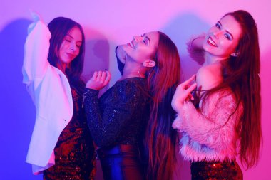 Üç güzel kız neon ışıkta dans ediyor. Parlak elbiseli kızlarla inanılmaz eğlenceli bir parti, bayramı kutluyorlar, gülümsüyorlar, gülüyorlar, dans ediyorlar, şarkı söylüyorlar. Gece kulübü dansı..