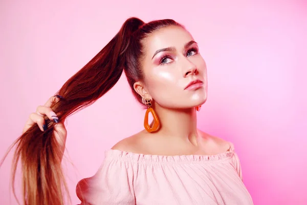 A beautiful girl, with long chic hair, a brunette holds well-groomed hair in a chicken. Chic girl with big earrings, accessories on a pink background.