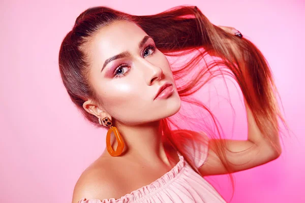 A beautiful girl, with long chic hair, a brunette holds well-groomed hair in a chicken. Chic girl with big earrings, accessories on a pink background.