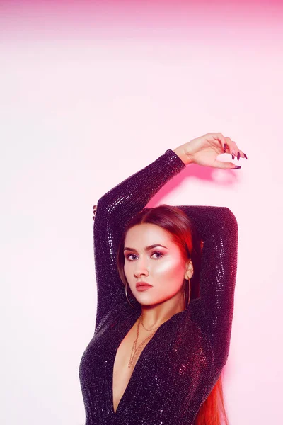 Parlak siyah elbiseli güzel bir esmer. Uzun saçlı kız, şık mücevherler, küpeler, kolye. Hassas pembe makyaj, kız dans ediyor. Pembe neon ışık
