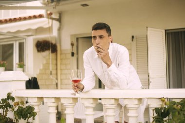 Bir adam terasta duruyor ve puro içiyor, kırmızı / pembe şarap içiyor. Zengin bir adam okyanustaki bir evde dinleniyor. Yalnız seyahat etmek, diye düşündü adam. Beyazlı bir adam tatilde..