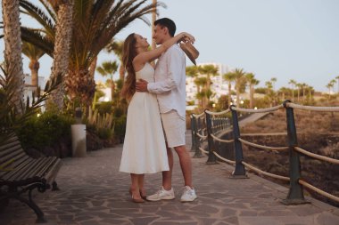 Okyanus kıyısında palmiye ağaçlarının arka planında gezinen bir çift. Beyaz randevulu bir adamla birlikte uzun beyaz elbiseli bir kız. Balayı, seyahat, turizm, Kanarya Adaları.